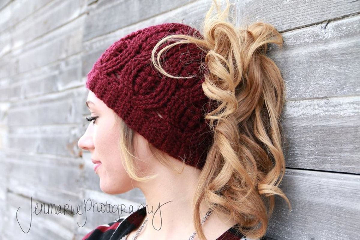 Blond woman side profile against gray wooden background in a maroon beanie with a blond ponytail sticking out. 