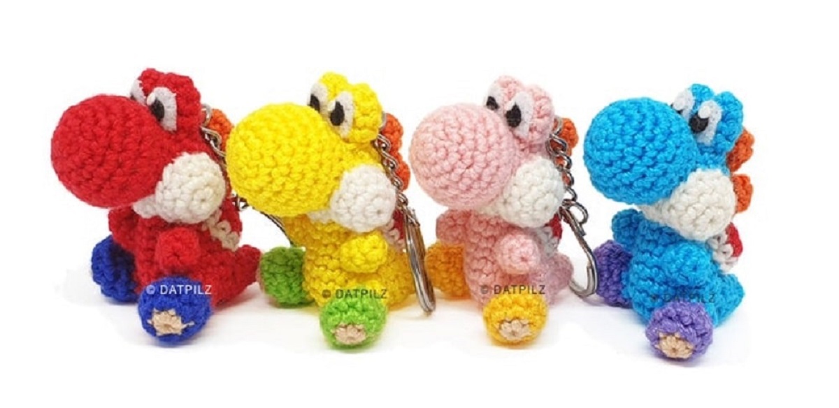Four small crochet Yoshi keyrings sitting in a row. There is a red, yellow, pink, and blue Yoshi all with yellow feet. 