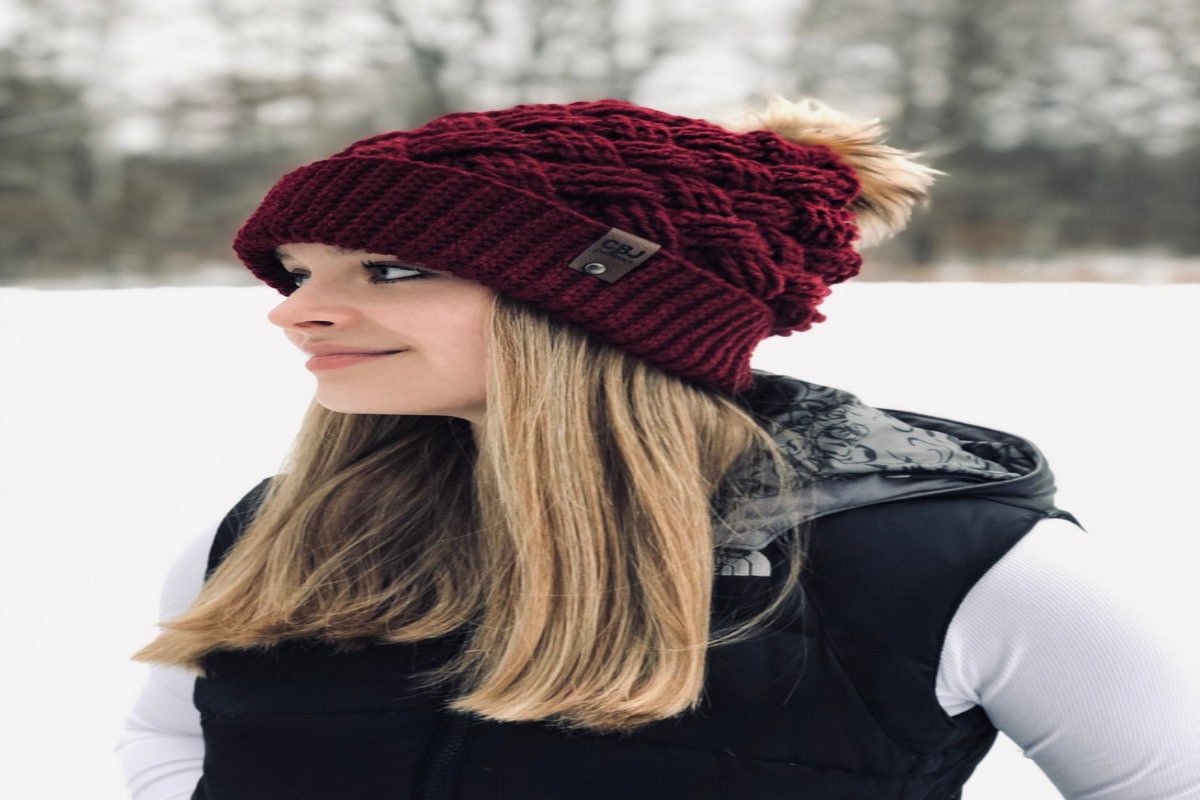 A blonde woman standing sideways by a snowy field wearing a dark red crochet hat with a fluffy bobble on top.