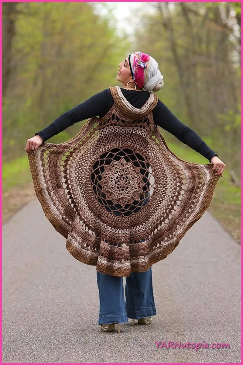 Woman standing on a path by some trees with a knee length brown and cream vest on, with a large mandala on the back.