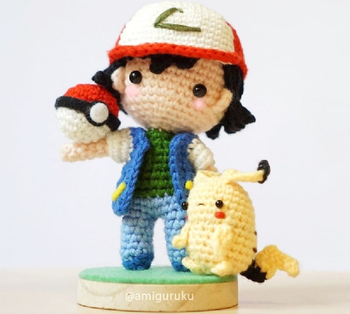 Stuffed crochet Pickachu standing next to a crochet Ash holding a large Pokeball in his hand and standing on a small grassy plaque.
