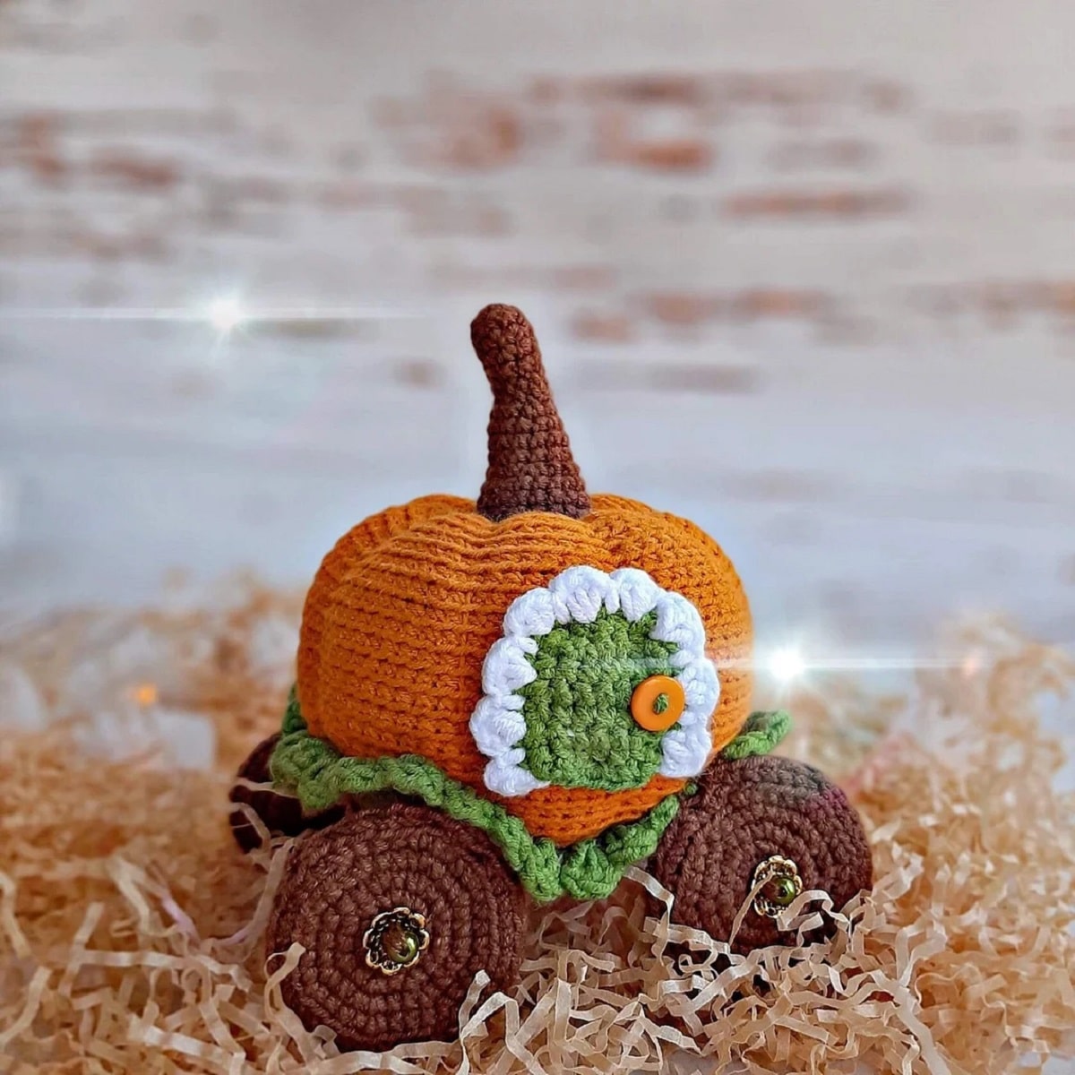 An orange crochet pumpkin carriage with a green door and brown wheels on the bottom on orange shredded paper.