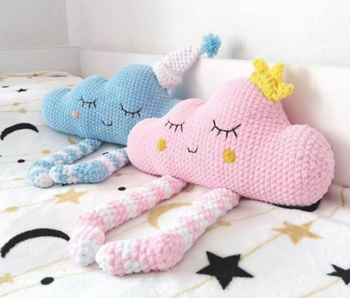 Two cloud shaped pink and blue crochet cushions with simple sleeping expressions and legs dangling from the cushions. 
