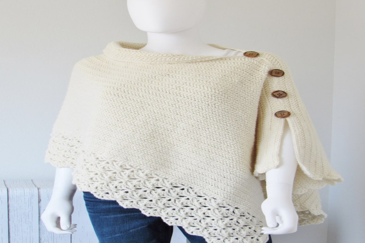 A cream poncho with a small slit and decorative chunky buttons on the right arm. A scalloped band with looser knits sits on the bottom.