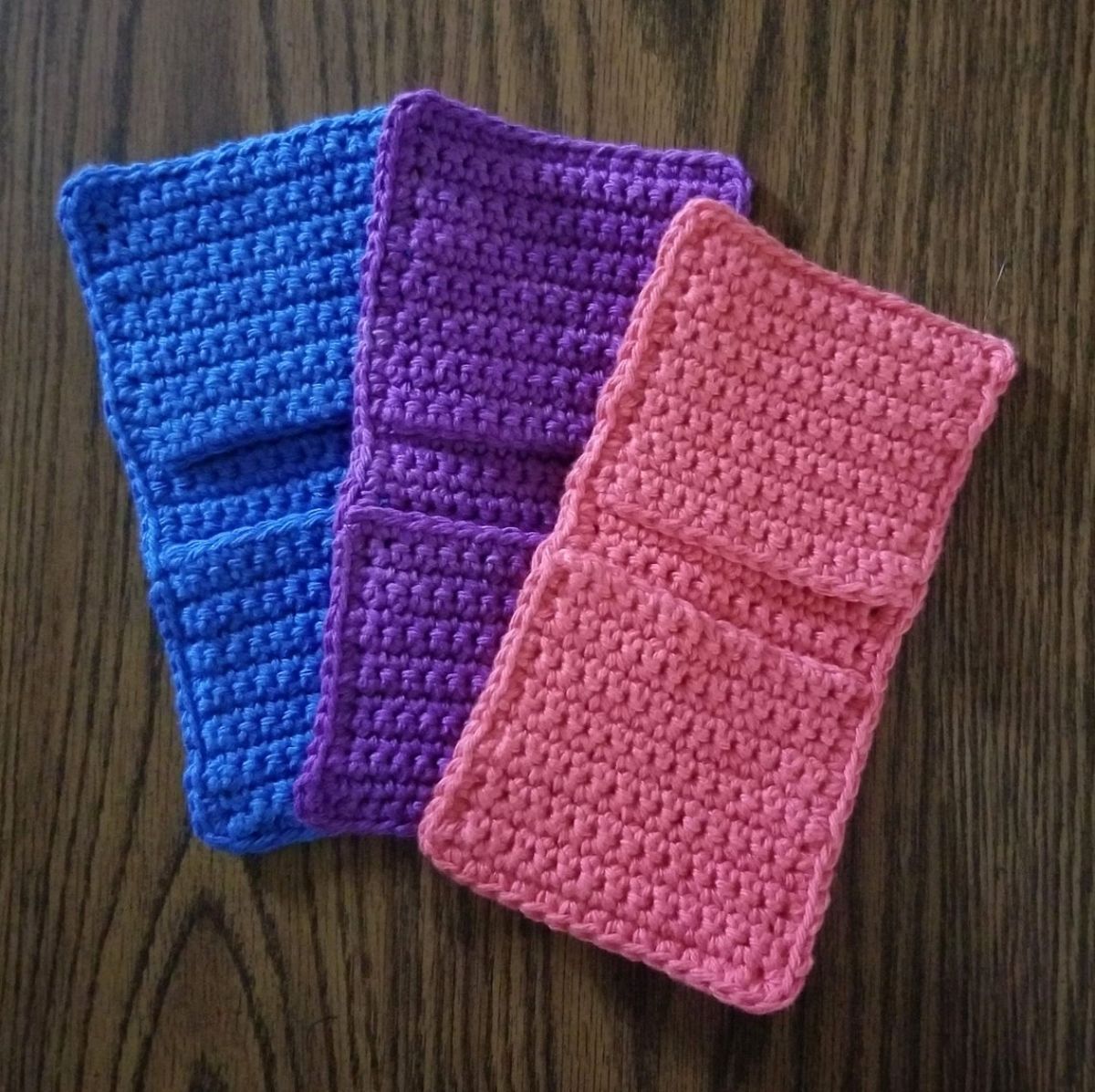 Rectangular potholders in pink, purple, and blue fanned out next to each other on a dark wooden table.