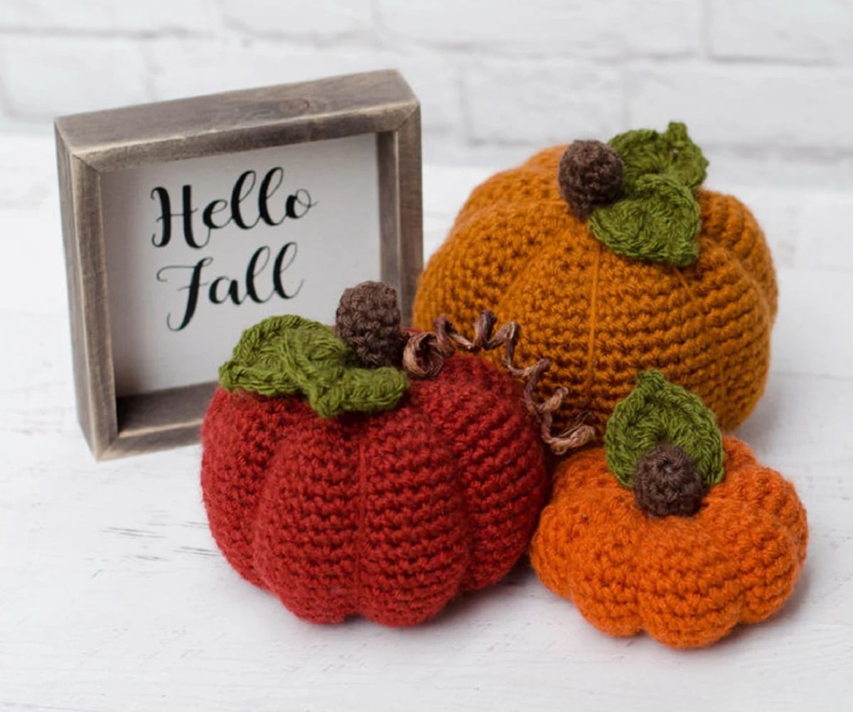A large orange crochet pumpkin with green leaves on the top next to smaller red and orange pumpkins with green leaves on the top. 