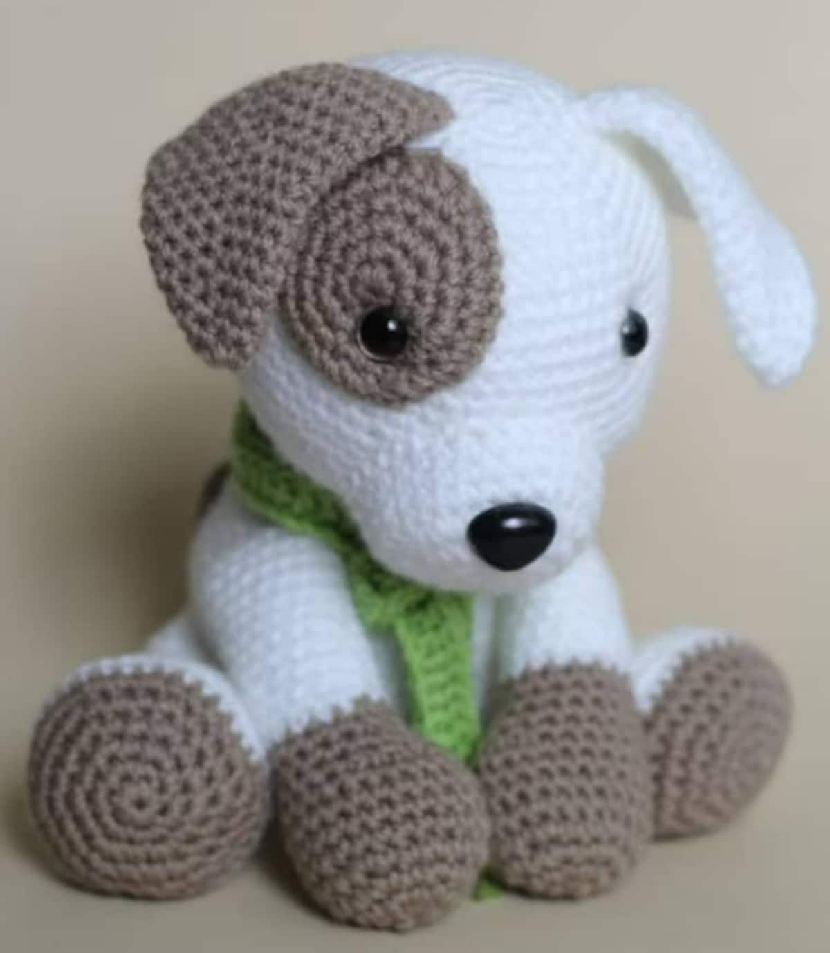 Crochet stuffed white dog with a brown patch over one eye, one brown ear, and brown feet wearing a green scarf.