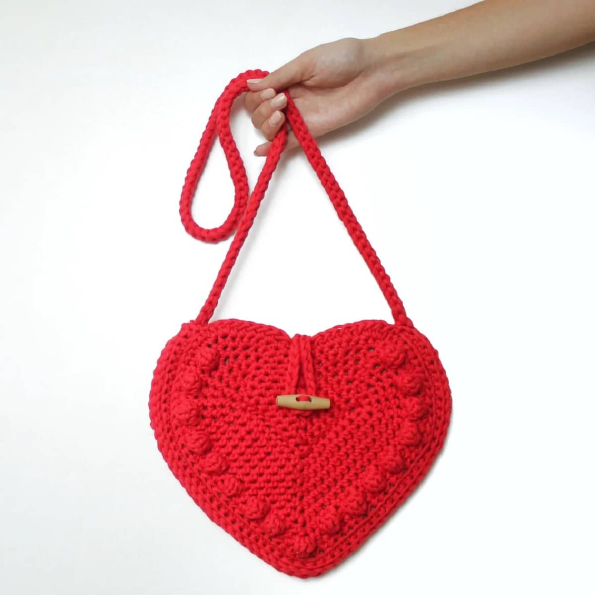 Red crochet heart shaped bag with bobbles stitched around the sides and a peg at the center to fasten dangled by its long strap by a hand.
