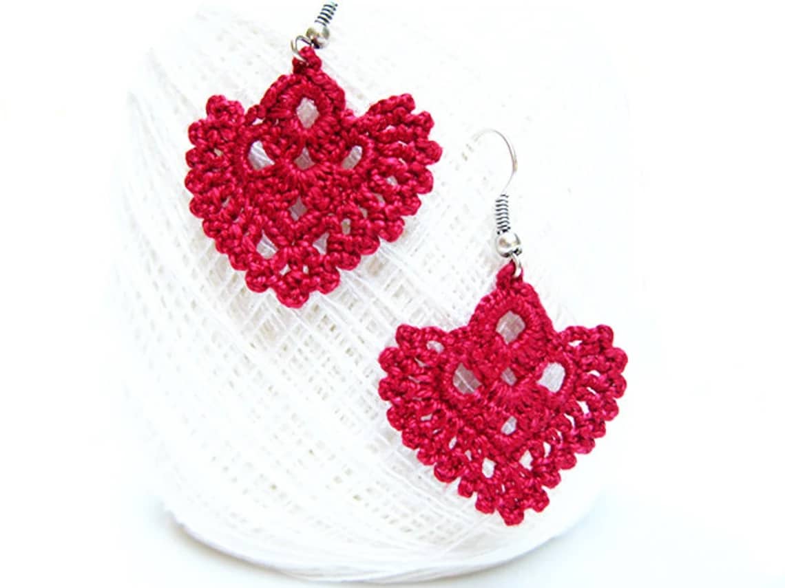 Two red crochet earrings with hearts in the center and a lace style trim dangling from a ball of white yarn.