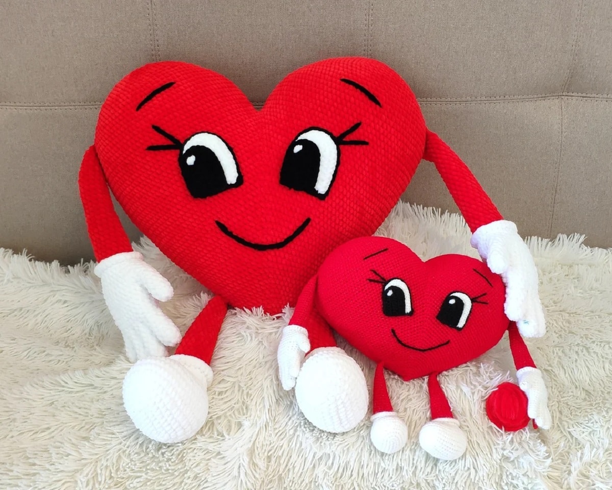 Large crochet red heart cushion with cartoon style eyes and smile, holding a mini version in the same style with red arms and legs with white hands and feet