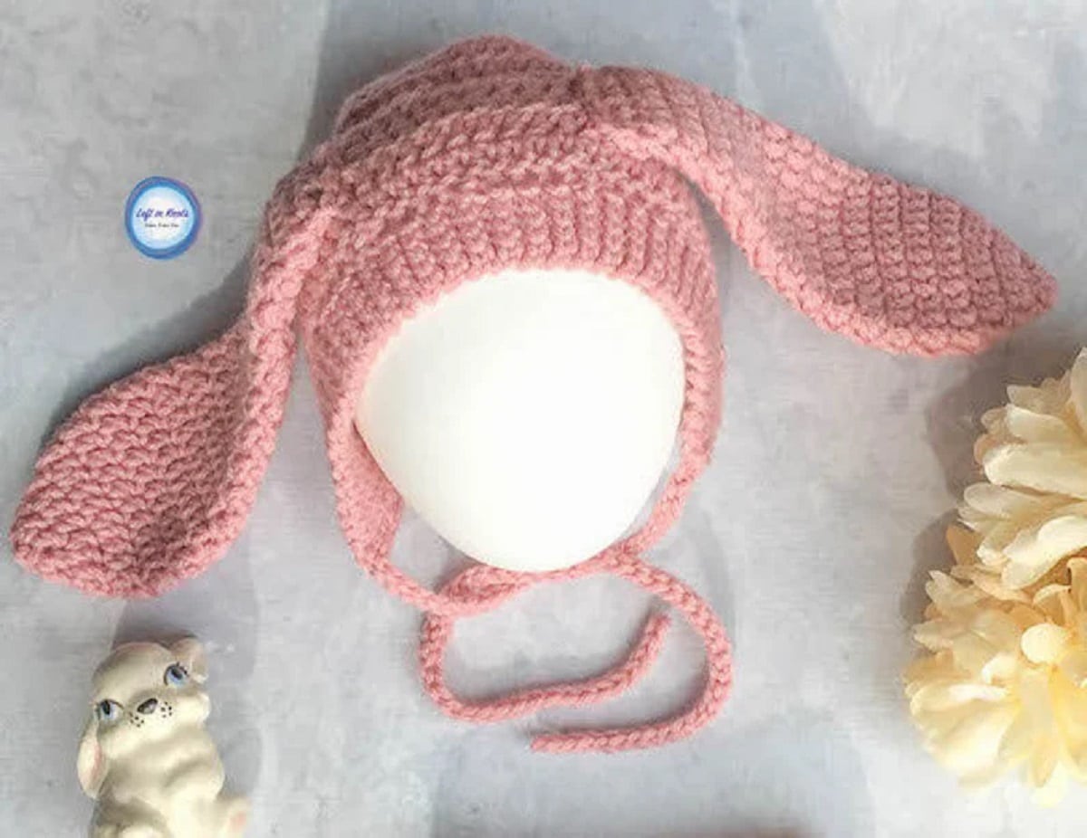 A pink crochet hat with large bunny ears on either side and pink braided straps to secure the hat.