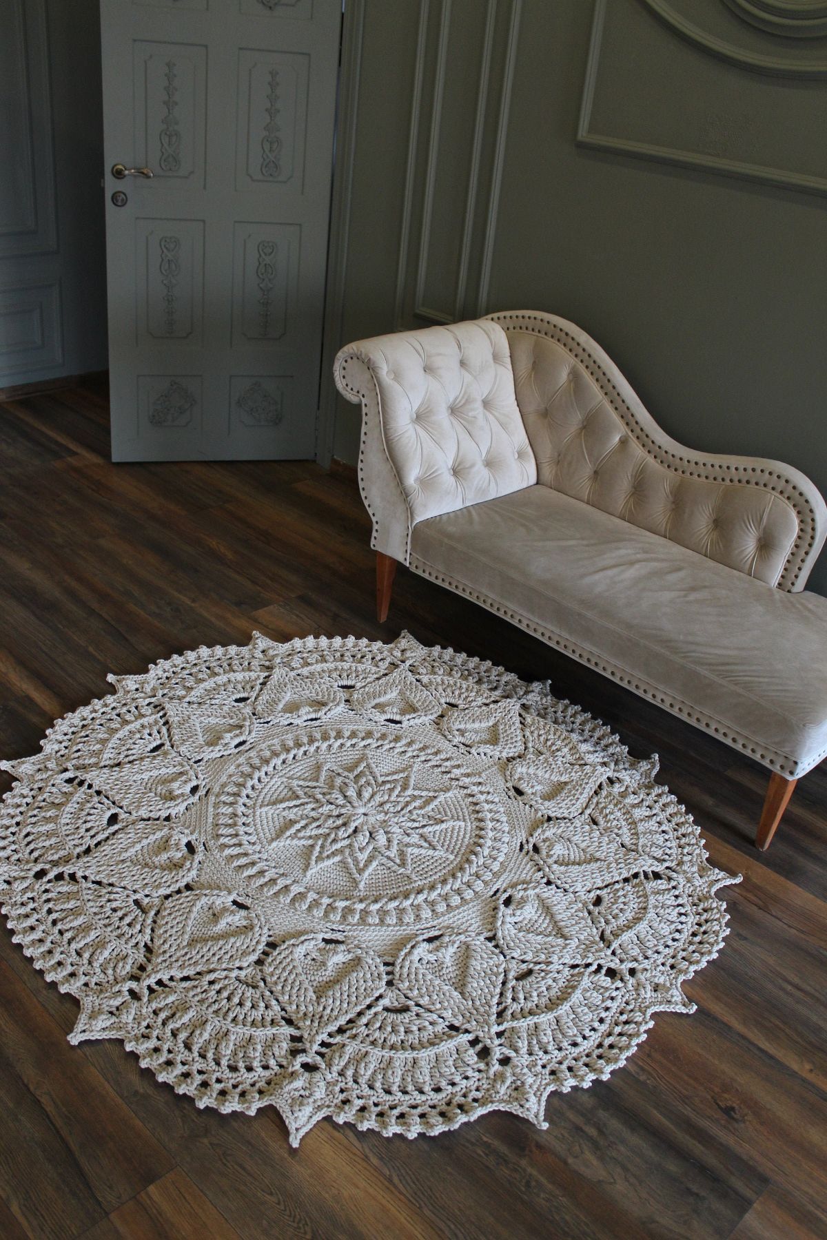 Large cream round rug with a doily outer edge and large petal-like pieces and a round center with a flower on wooden flooring next to a cream sofa.