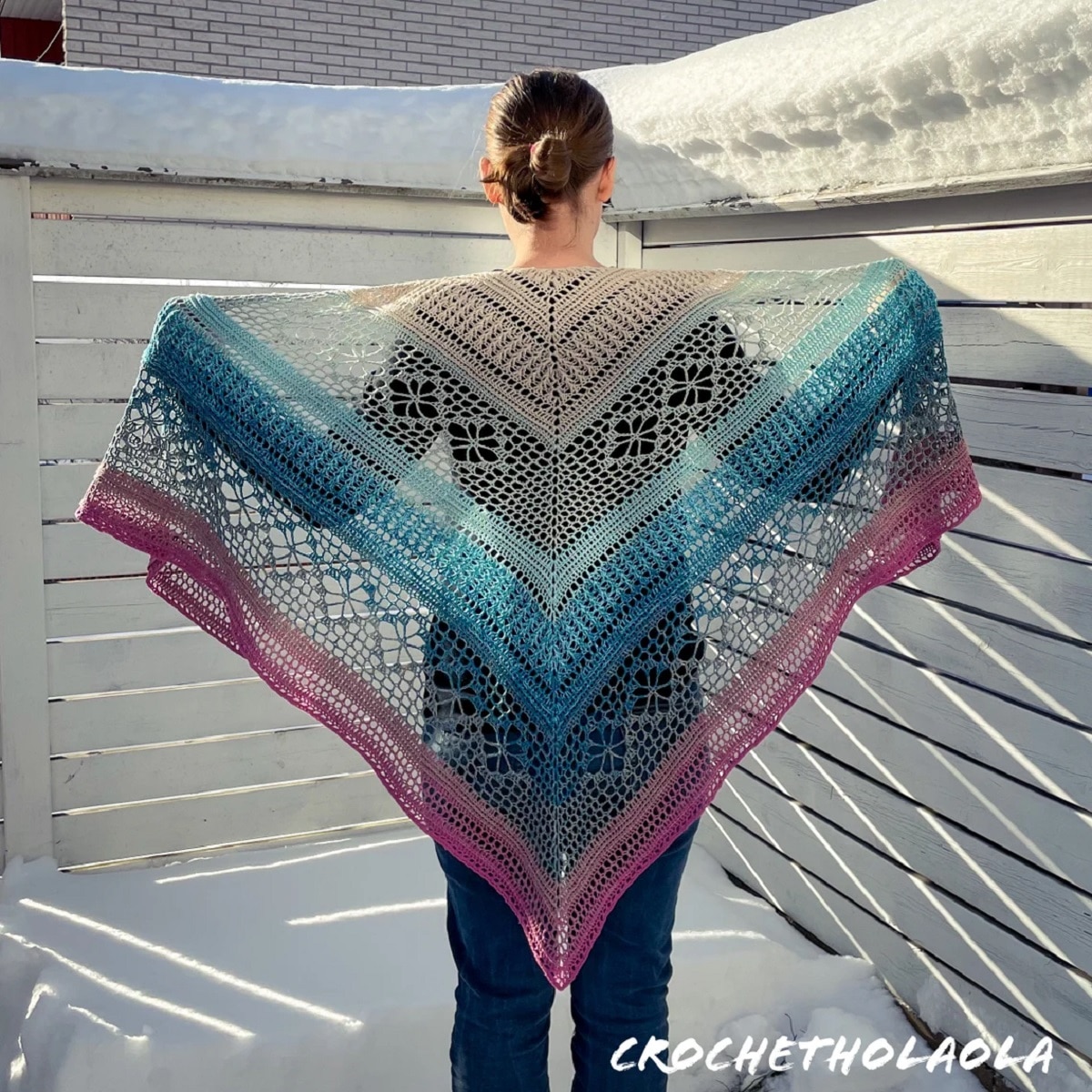 The back of a woman holding a pink, blue, and cream crochet shawl with horizontal straps and diamond shapes in the cream sections.