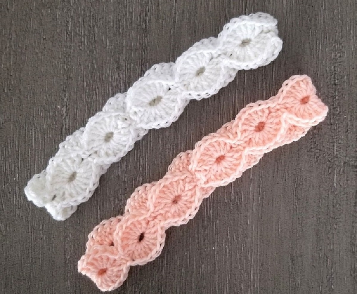 A salmon pink and white crochet headband featuring an open shell design on a gray wooden background.