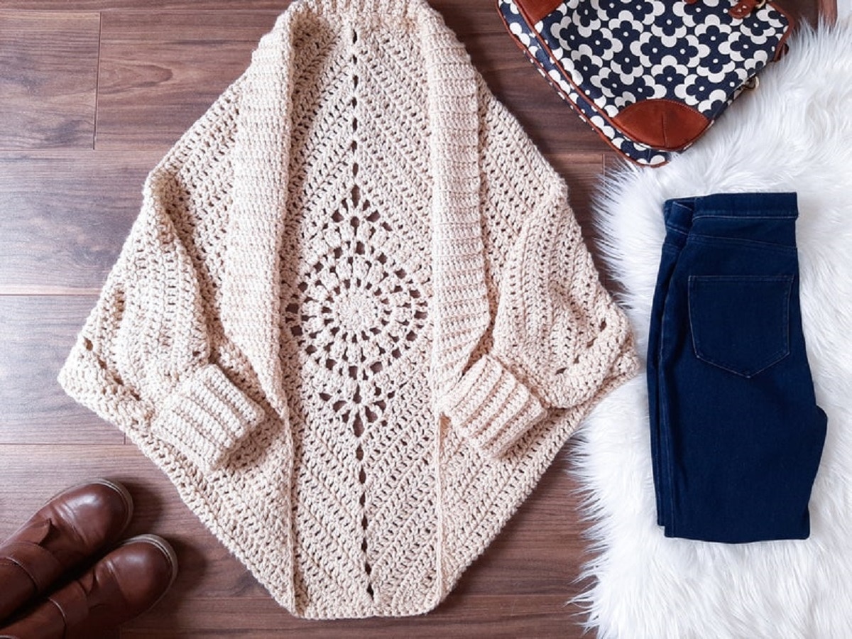 A cream crochet shrug with long cuffed sleeves and a small round mandala on the back on a wooden floor.