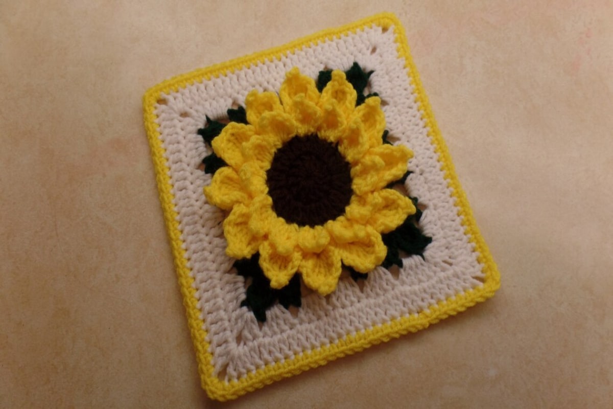 White crochet square with a yellow trim and 3D effect sunflower in the center. The sunflower has yellow petals and a brown center.