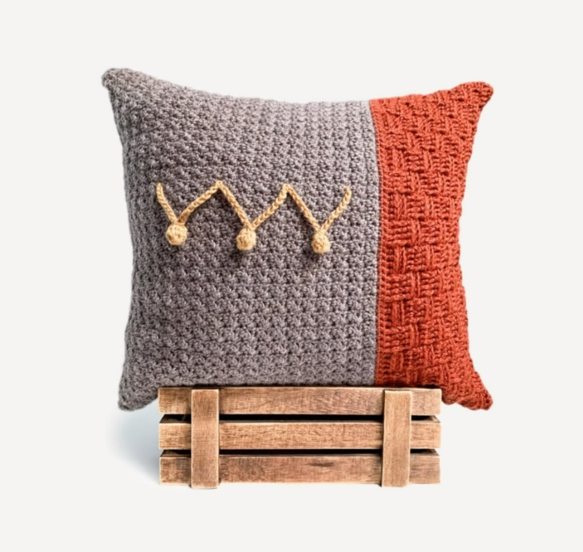 Gray crochet cushion sitting on a wooden box with one large red stripe and three golden bobbles with simple gold stitching in the center.