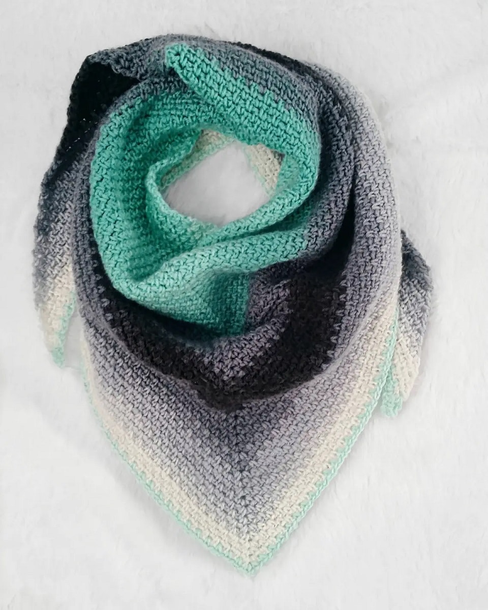 A white, gray, black, and green crochet triangle scarf wrapped into a circle on a white background.