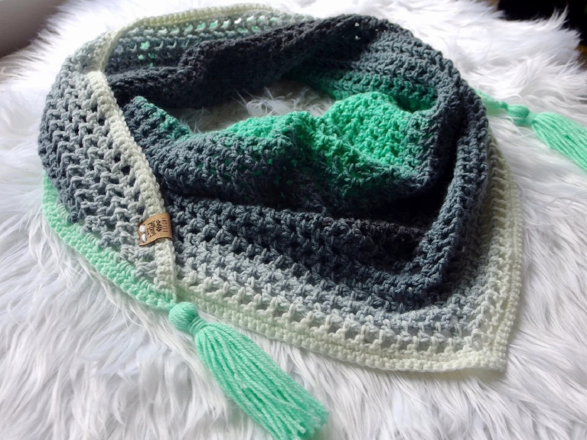 A gray, navy, and green crochet triangle scarf with two long green tassels folded on a white blanket.
