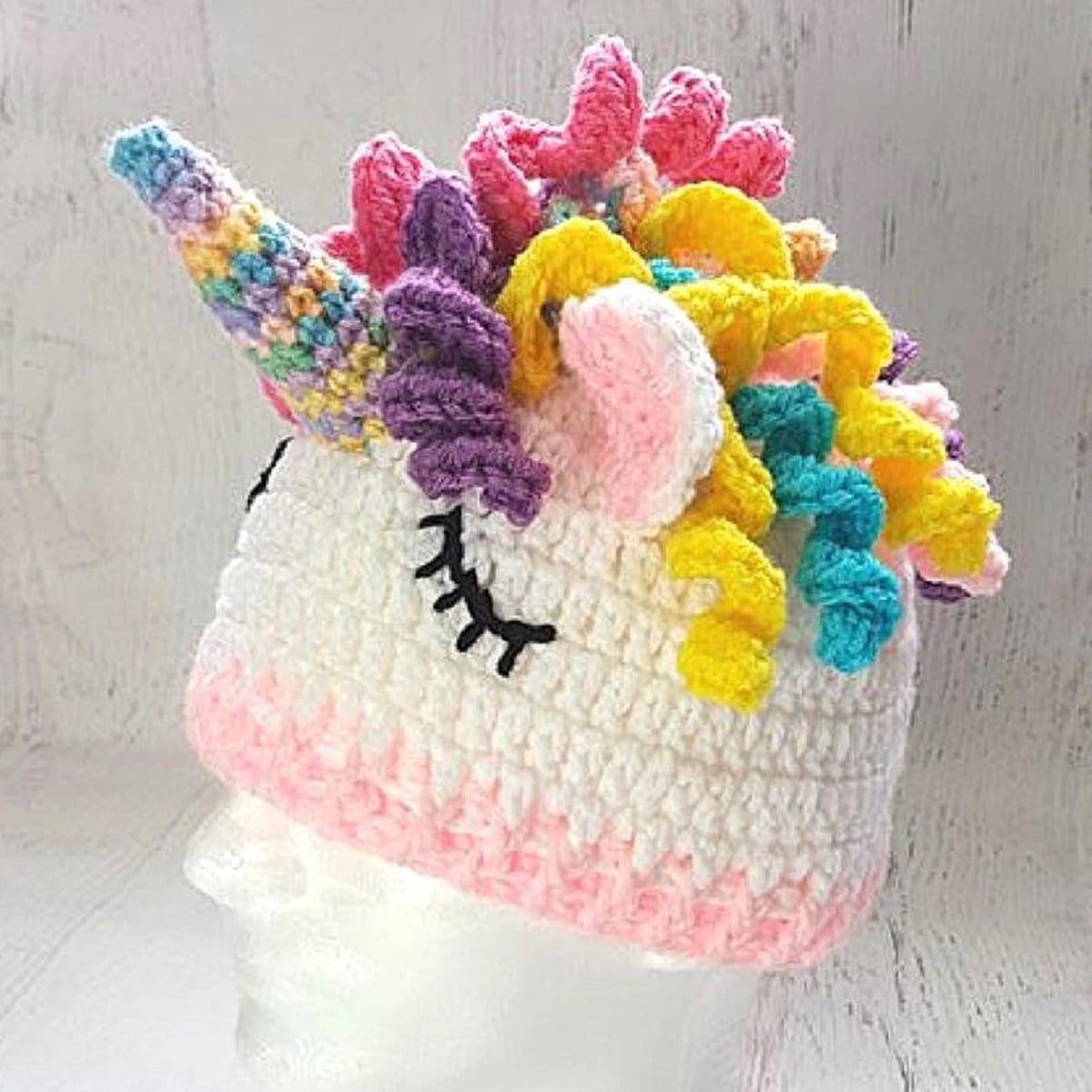 Mannequin wearing a white crochet beanie hat with closed eyes, a multicolored horn, and curled blue, yellow, purple, and pink hair on top.