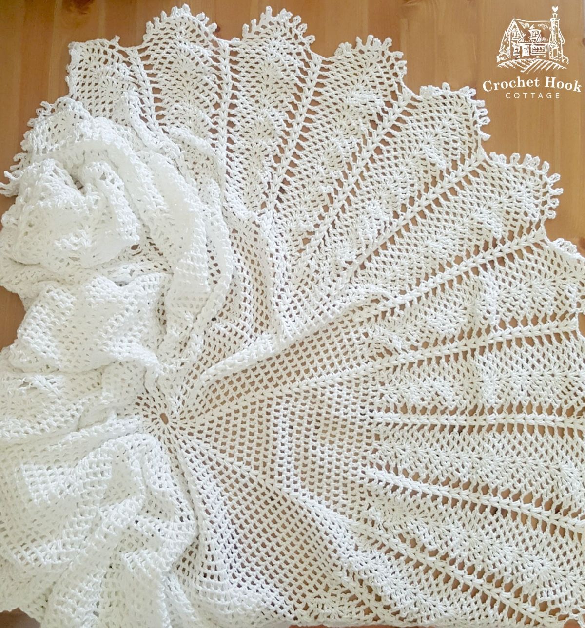 A white crochet baby shawl shaped like a fan spread out on a wooden floor.
