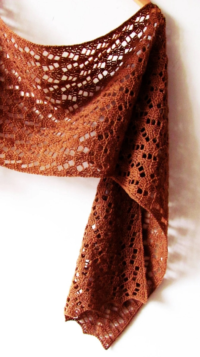 A brown lace crochet shawl hanging from a hanger on a white wall.