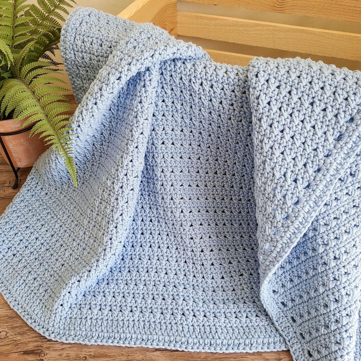 A blue crochet baby blanket spilling out of a wooden box next to a potted plant.