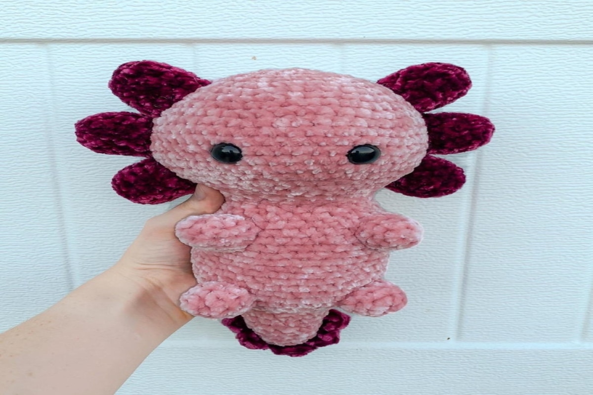 Pale pink crochet Axolotl with dark pink ears held in a hand by a white wall.