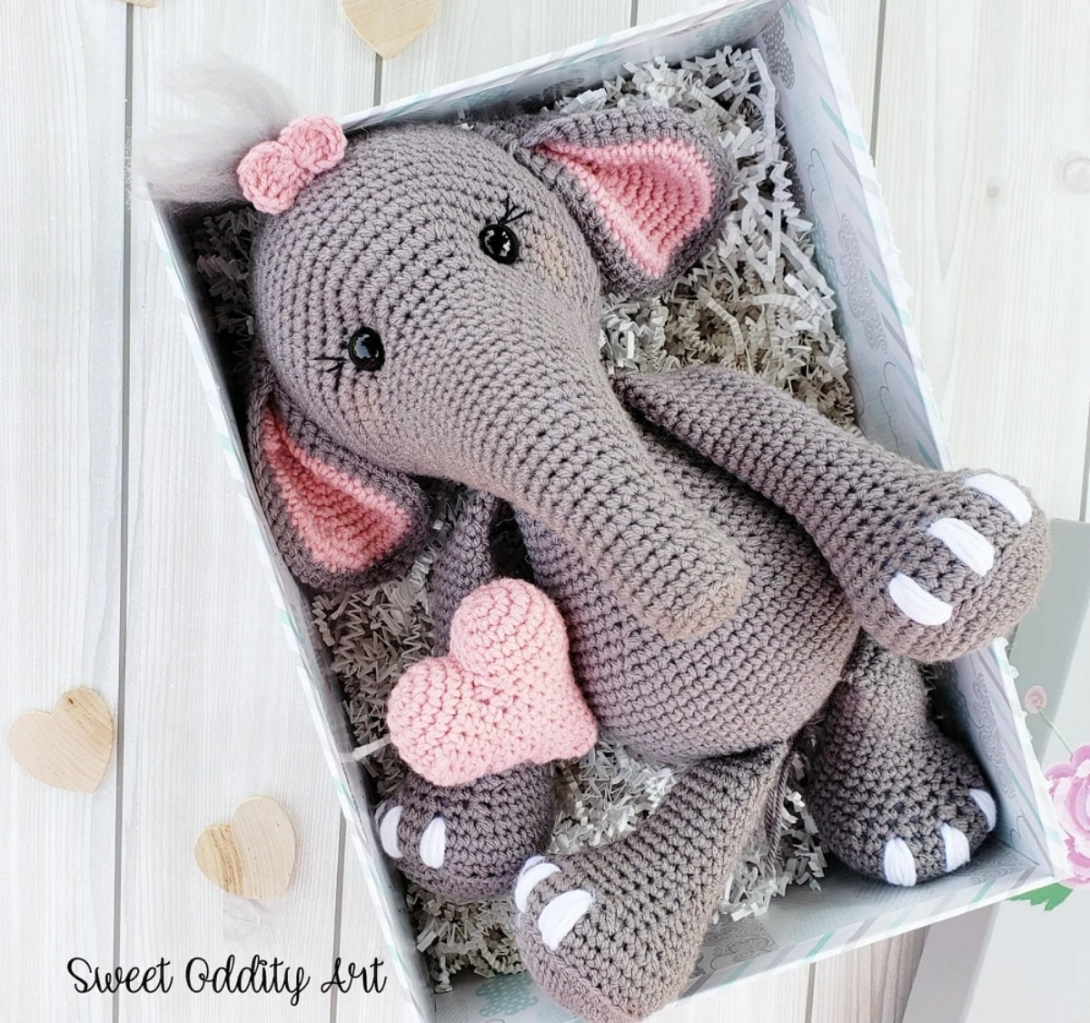 Large crochet gray Elephant with small pink ears and a pink bow on her head lying in a white box with a pink crochet heart next to her.