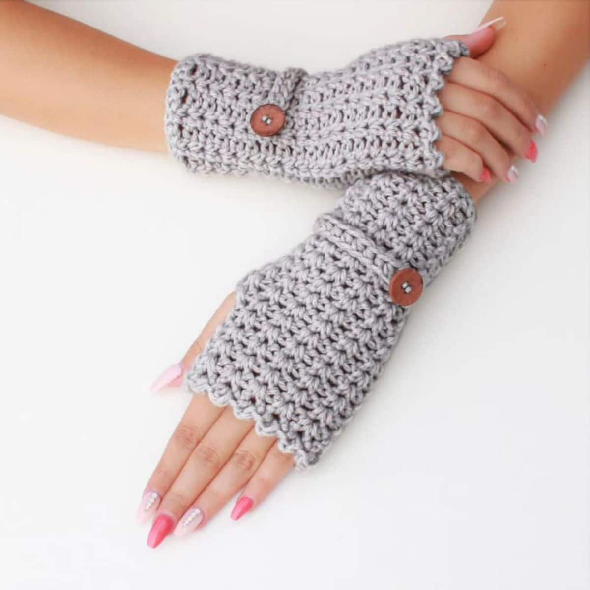 Manicured hands wearing gray crochet fingerless gloves with a band and button around the wrist to secure them.