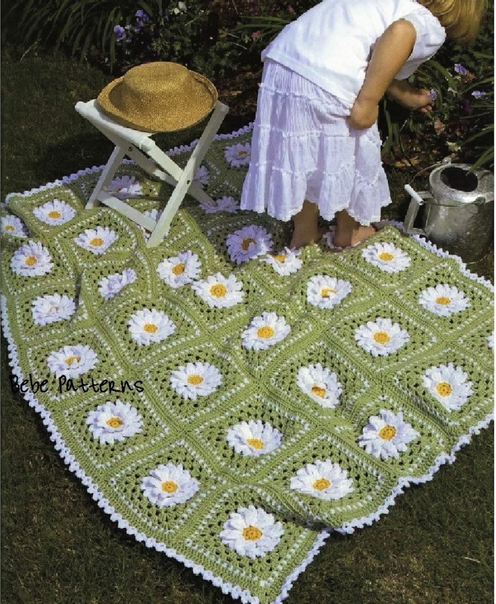 Small child in white bent over next to some plants standing on a green crochet Afghan with white daisies in squares and a white narrow trim on all sides.