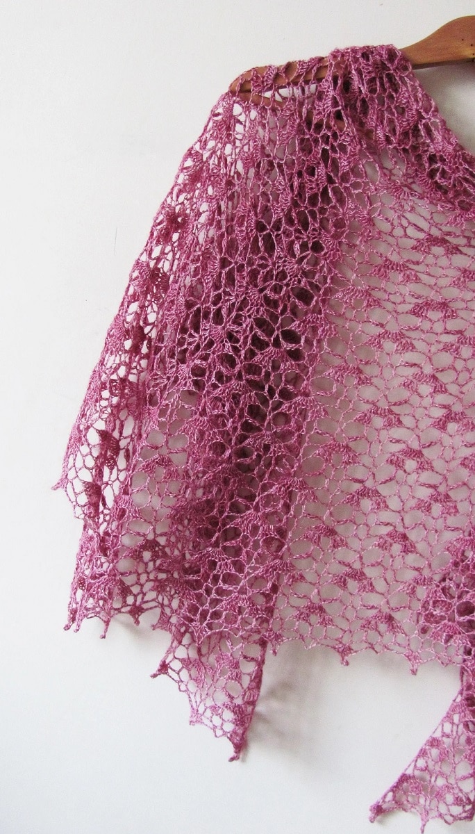 Half of a pink crochet loosely weaved crochet shawl hanging on a wooden hanger against a white wall.