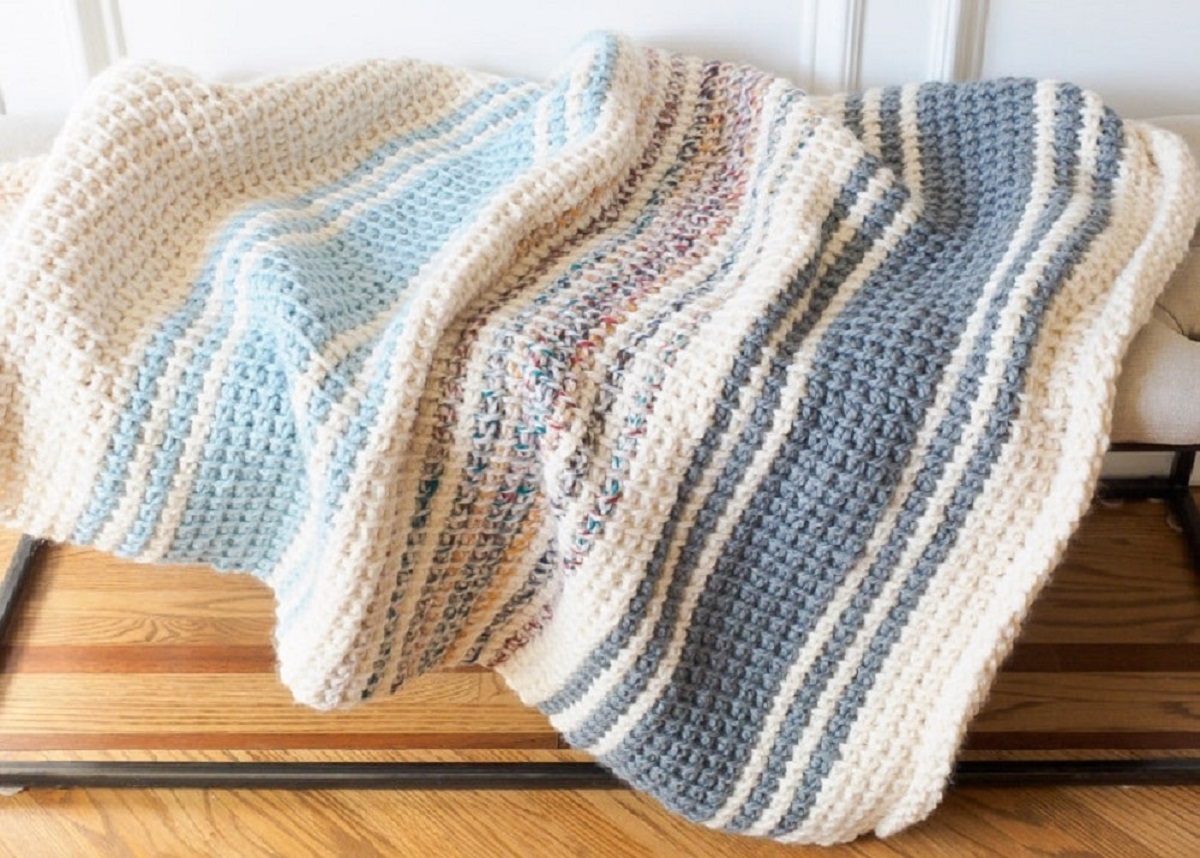 A white, blue, pink, and gray striped crochet blanket bunched up and folded on a wooden floor.