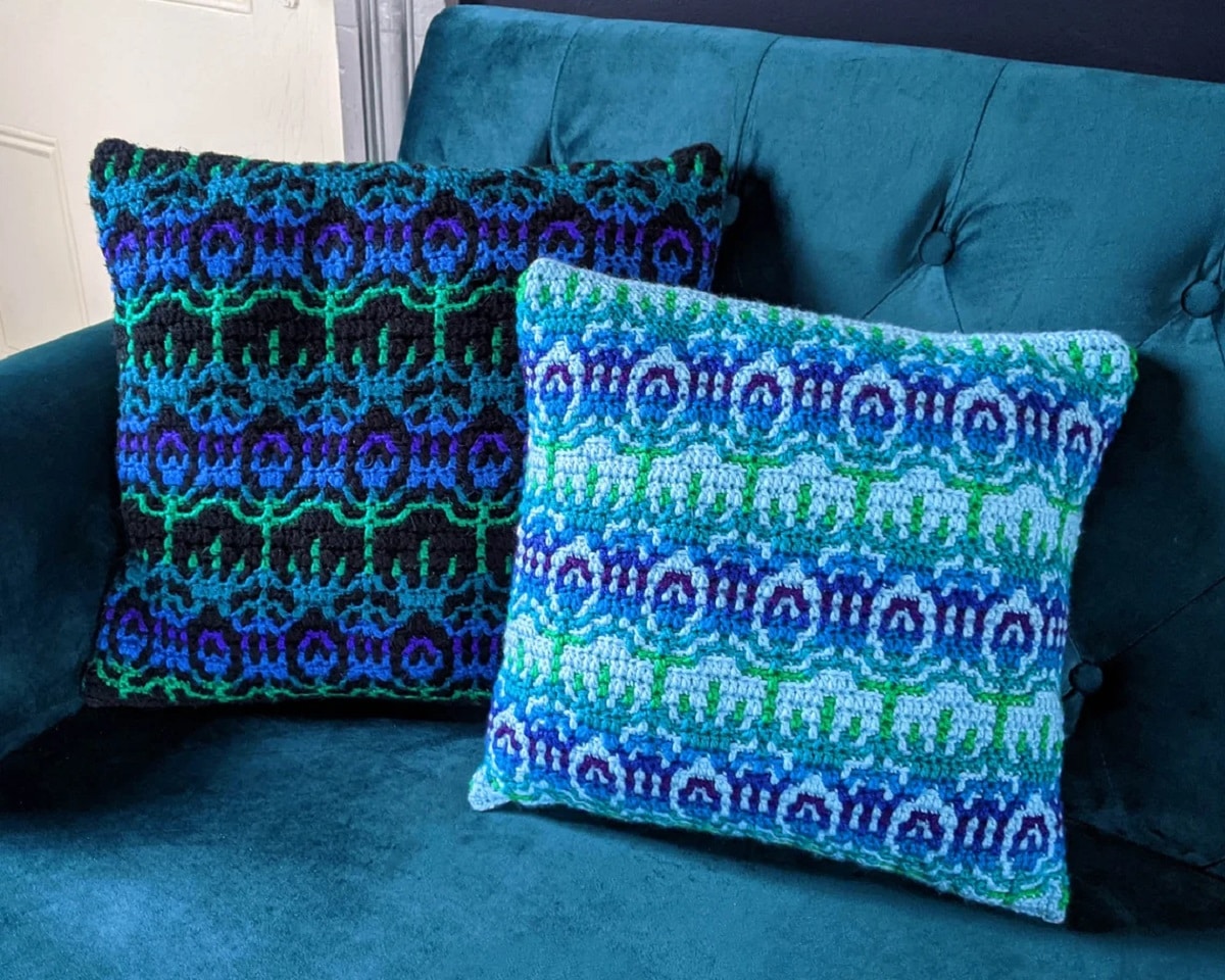 A dark blue, green, purple, and black peacock pattern crochet cushion next to a light blue, green, and purple cushion with the same pattern.