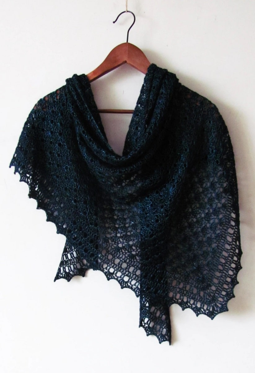  Dark blue crochet shawl with a small lace trim on all sides wrapped around a wooden hanger on a white background.