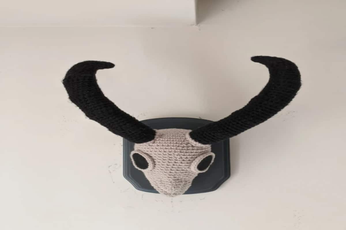 A white crochet goat skull with black eyes and long black horns attached to a gray mount on a white wall.