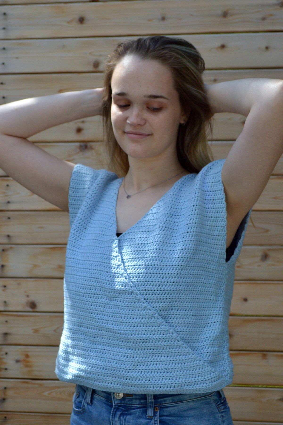 A young brunette woman with her hands behind her head in a v-neck crochet blue vest against a wooden background.