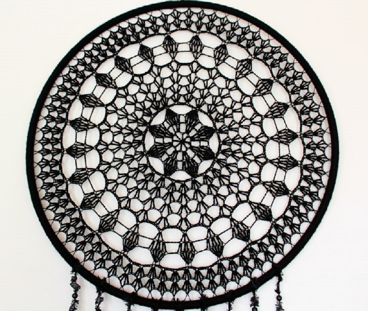 Large black dream catcher hanging on a wall with intricate crochet designs and black strings hanging from it.