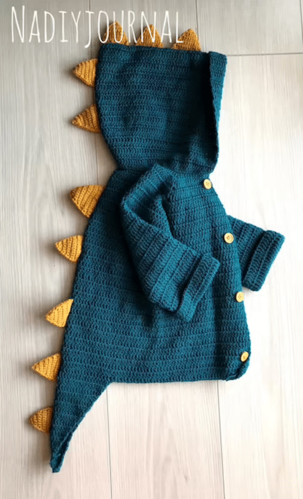 Small teal crochet cardigan with long sleeves, buttons down the center and yellow dinosaur spikes down the hood, back, and tail at the bottom.