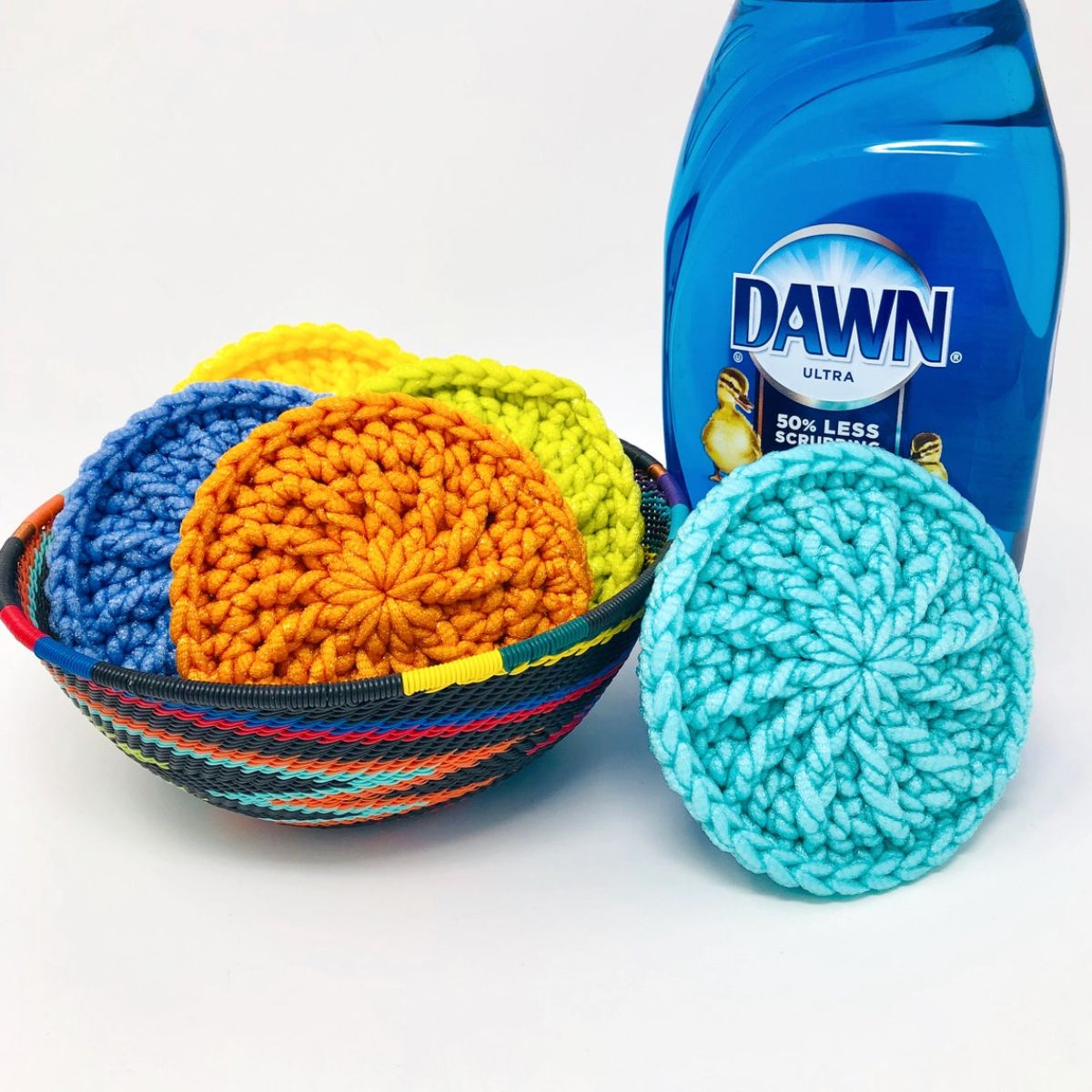 A basket filled with orange, green, yellow, and blue round crochet scrubbies next to an aqua scrubbie and a bottle of dish soap.