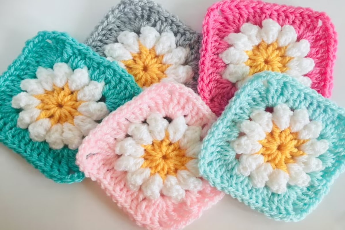 Five crochet granny squares with daisies stitched onto the center. Squares come in blue, pale pink, pink, green, and gray. 