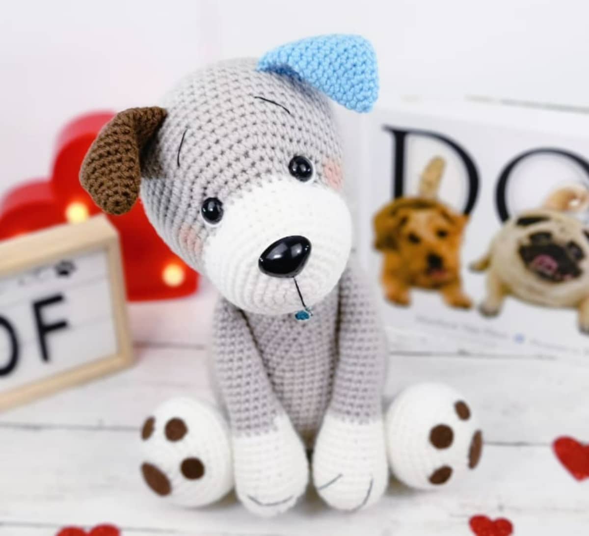 Light gray stuffed crochet dog with one brown ear and one blue ear sitting on a light wooden floor.