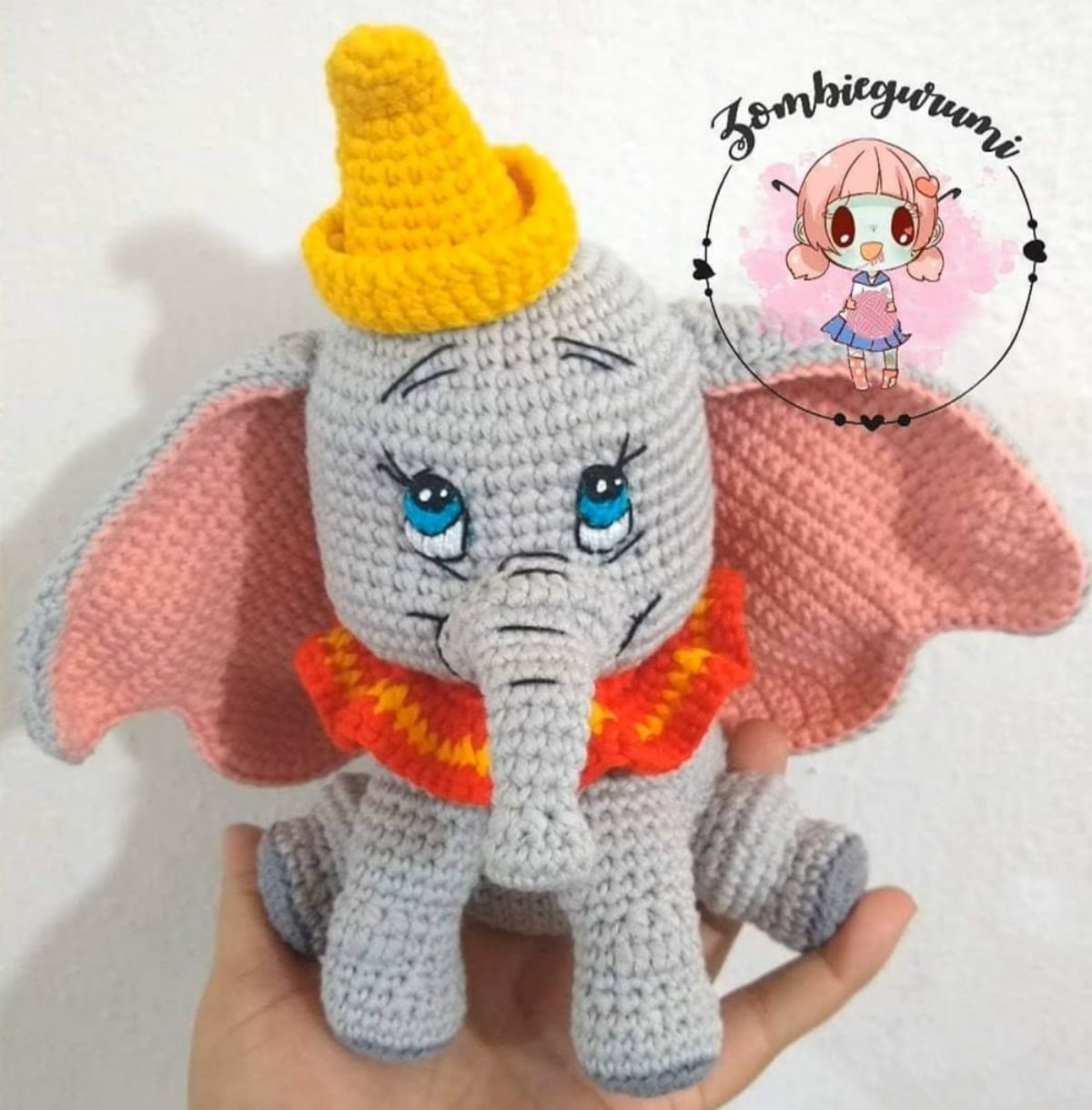 A hand holding a gray crochet Dumbo with large pink ears, a small yellow hat, and pink and orange ruffle around its neck.
