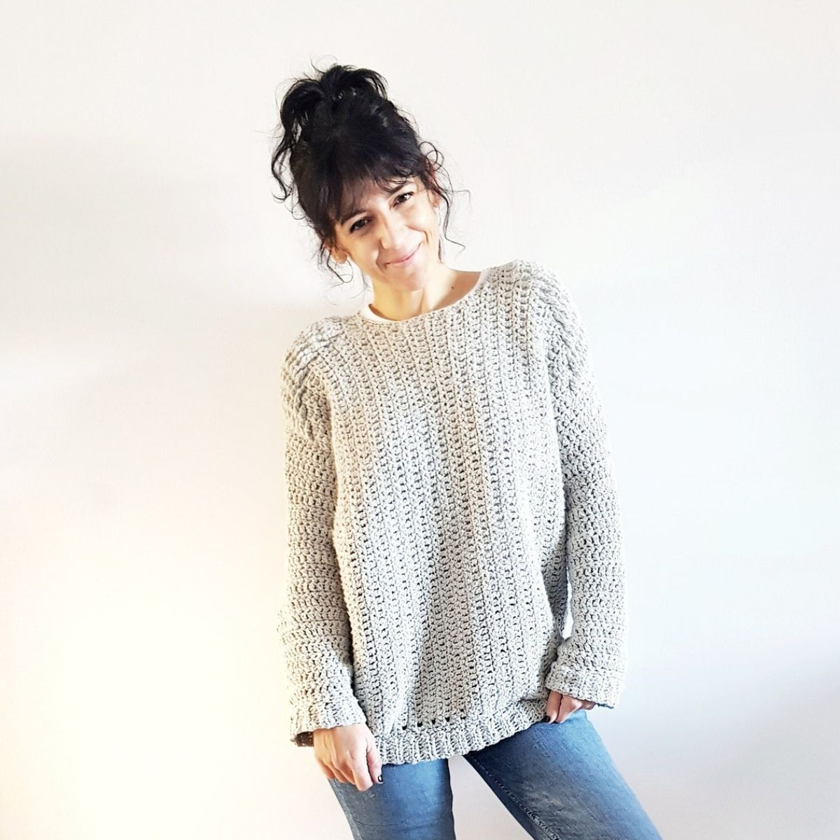 Smiling brunette woman in a long sleeved light gray crochet jumper with a chunky trim on the bottom against a white background.