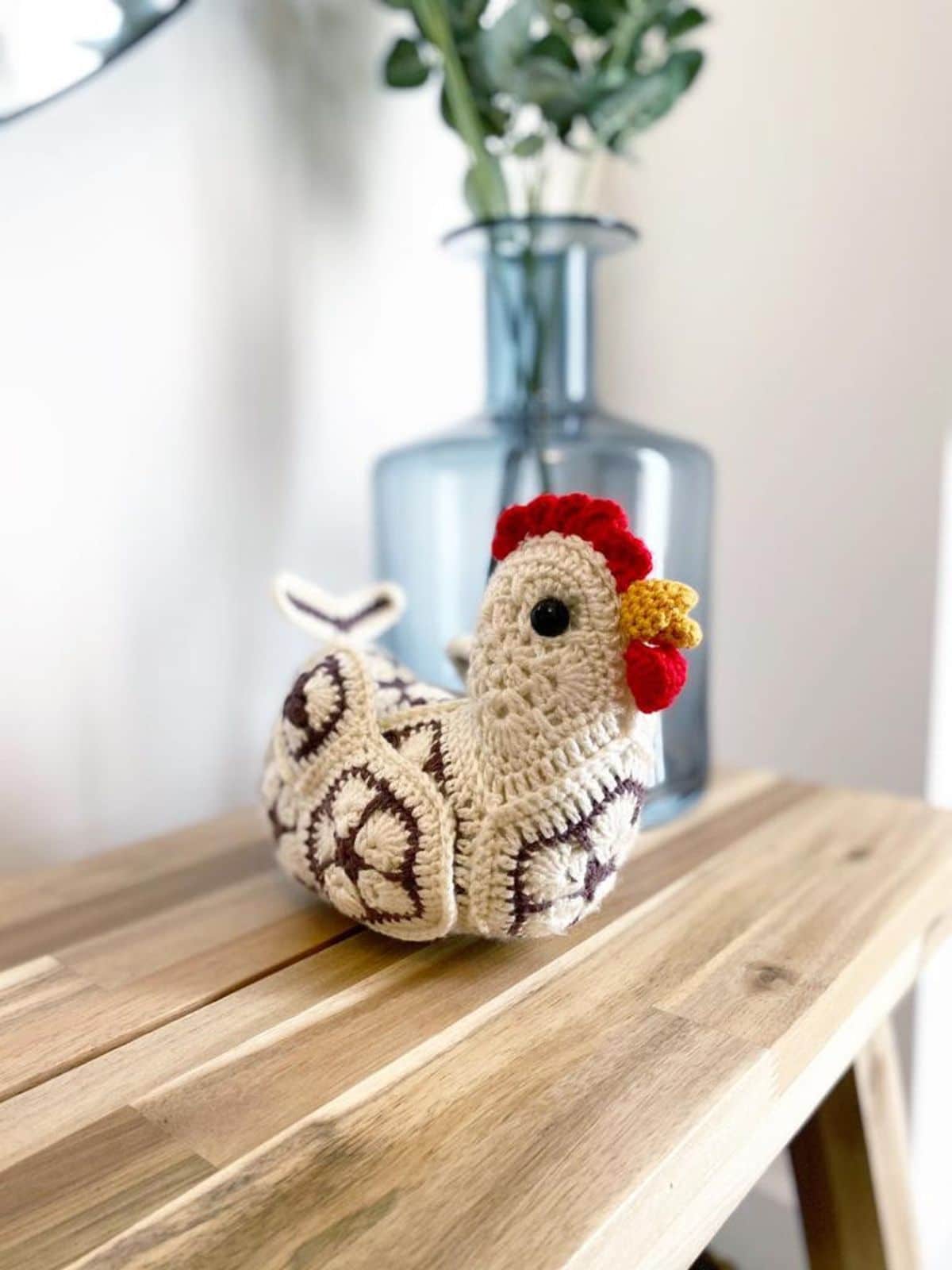 Small cream crochet Chicken with hexagons of brown flowers stitched around its body, a yellow beak, and red comb on a wooden table.