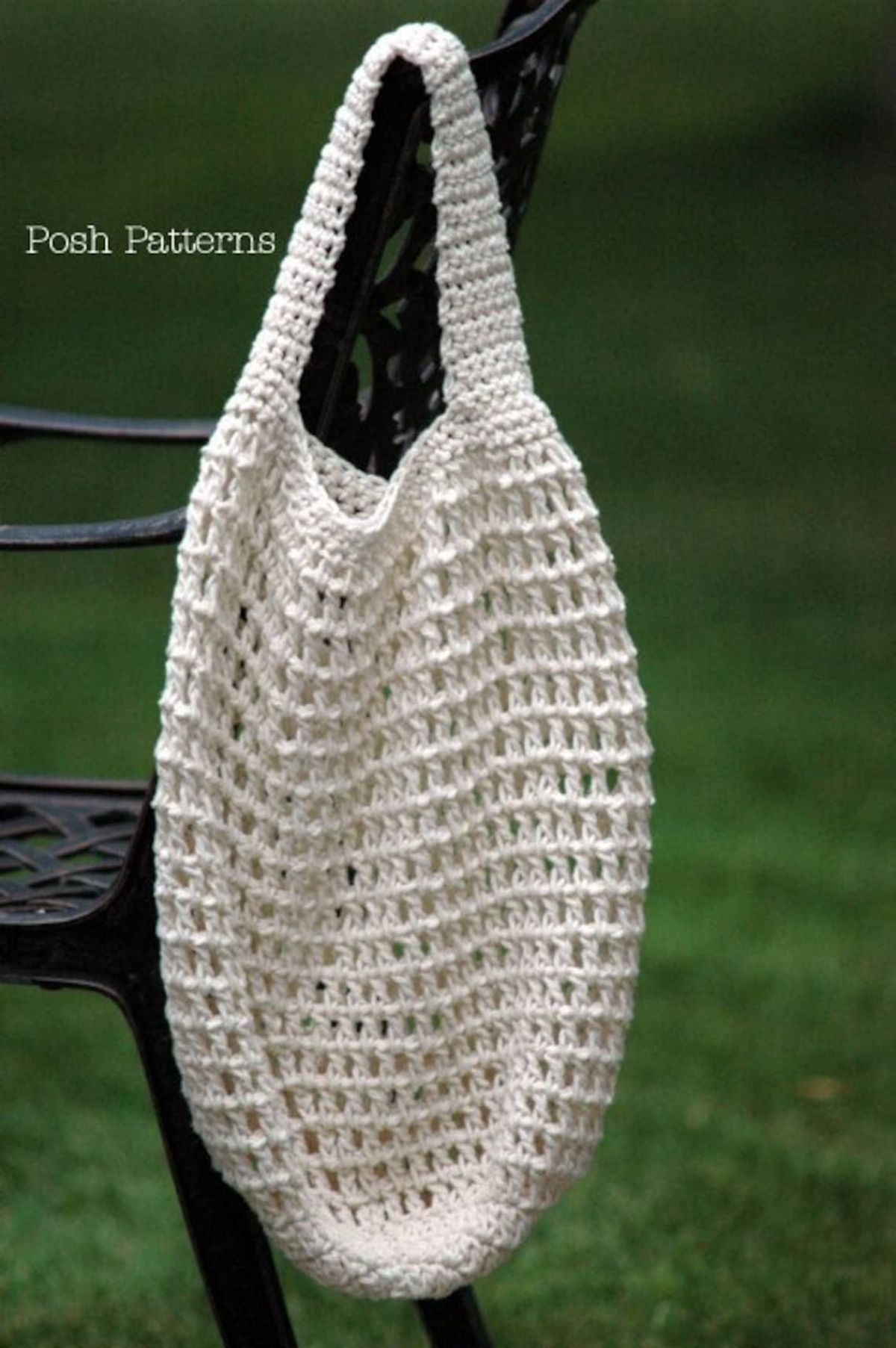 White crochet market bag with one thick shoulder strap hanging on the back of a black chair by some grass. 