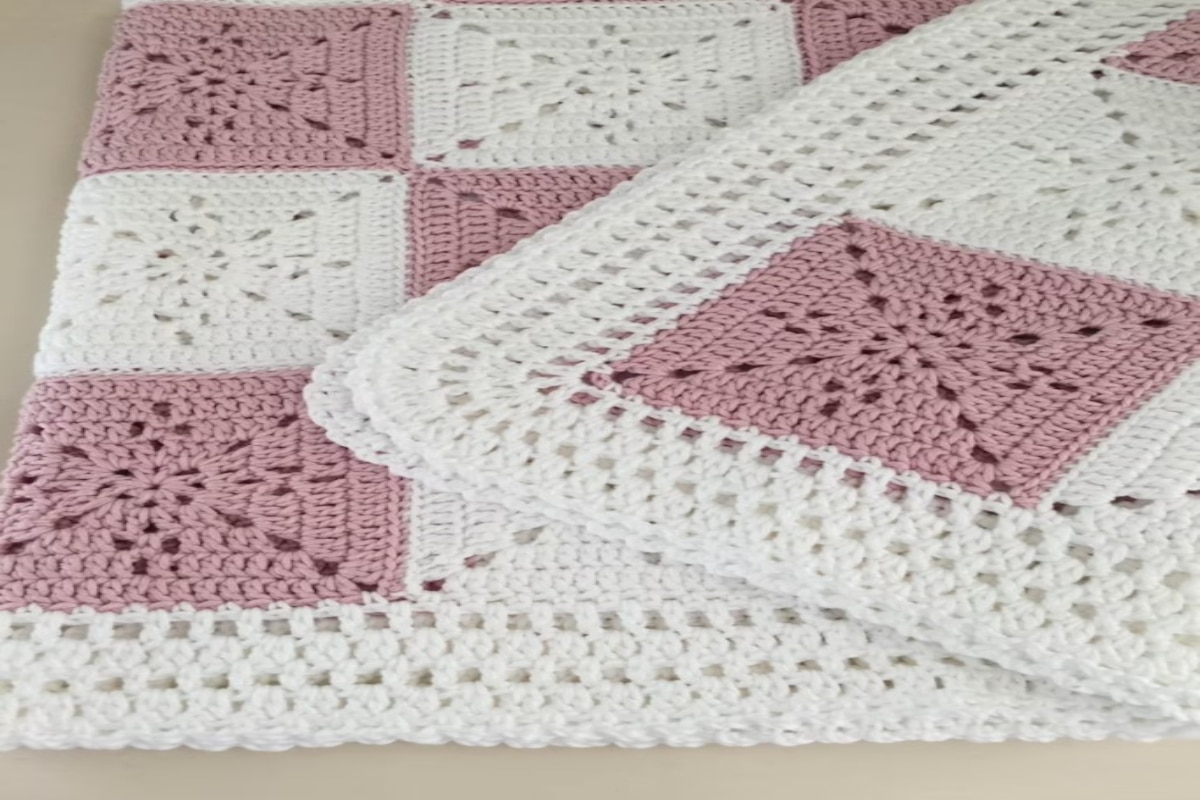 Granny square crochet blanket with pale pink and white squares and a white lace style trim around the folded blanket.