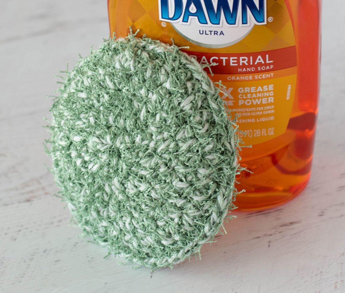 A round green crochet scrubbie balanced on an orange bottle of Dawn dish soap on a white counter.