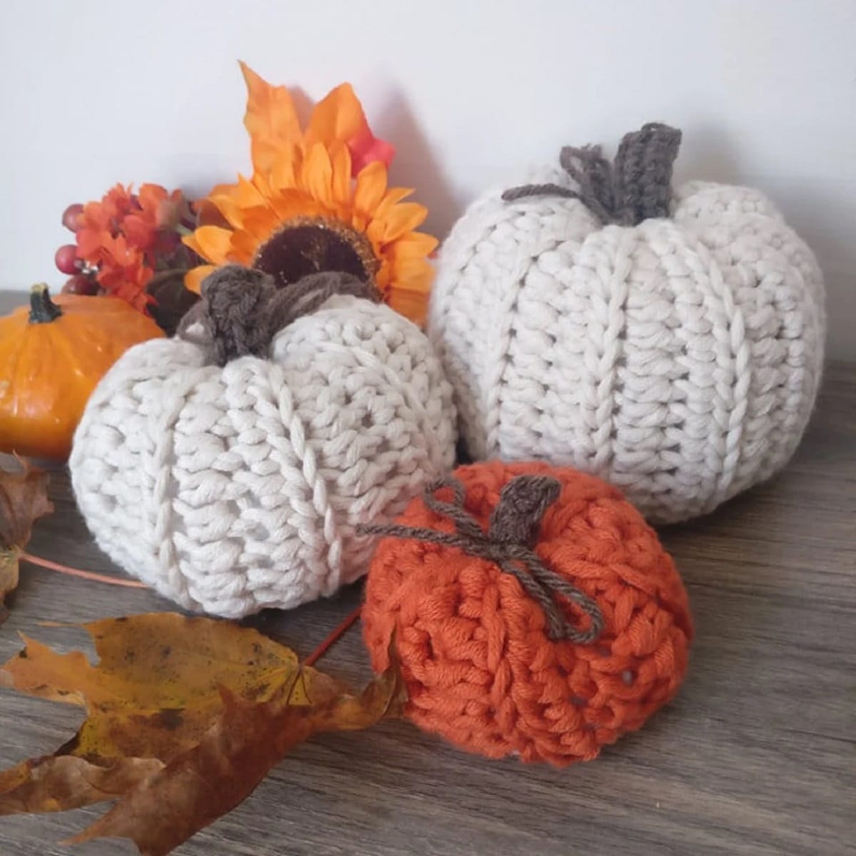 A small orange crochet pumpkin with a brown bow on top in front of two larger white crochet pumpkins on a wooden table.