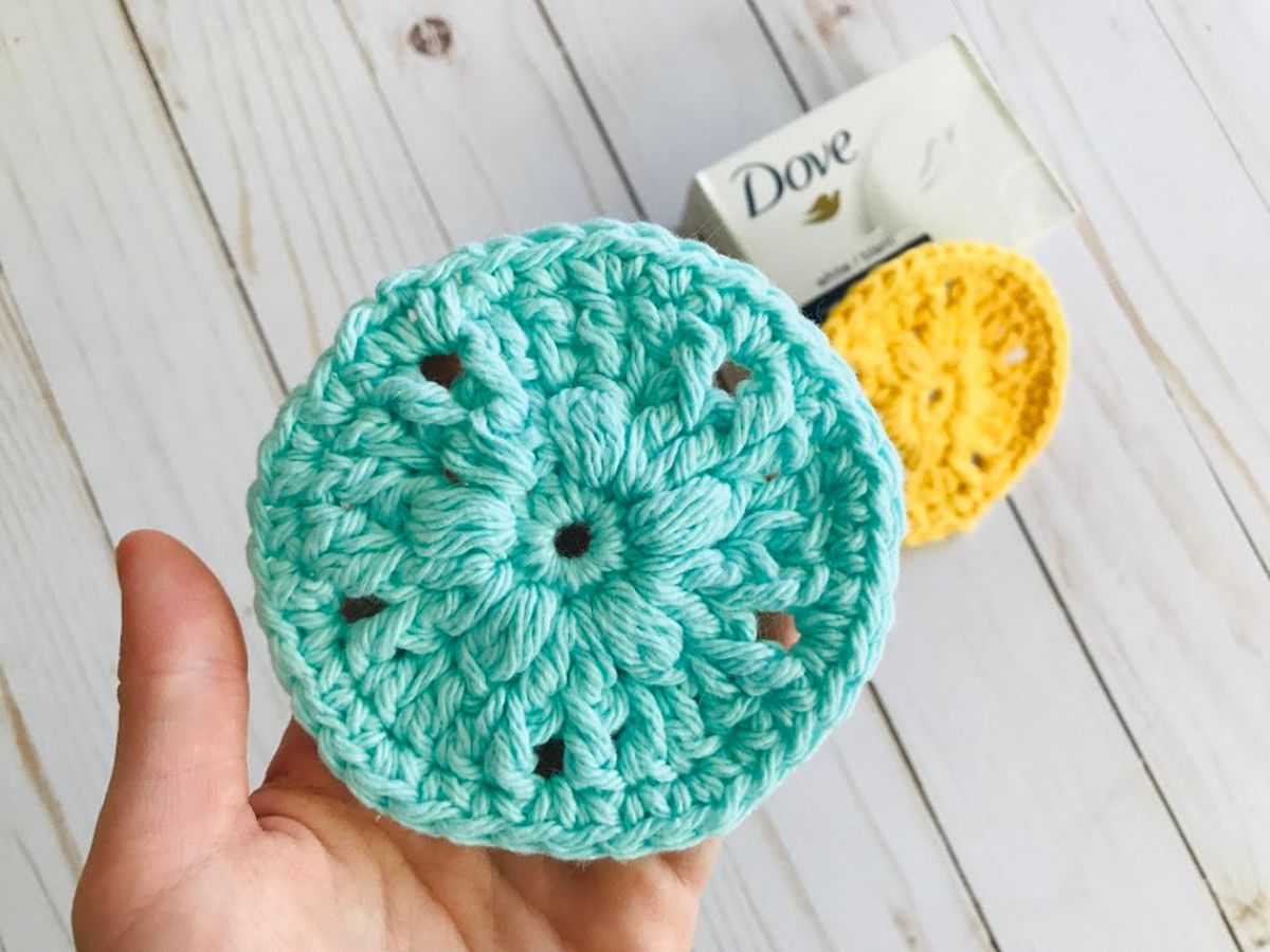 A hand holding a blue round crochet scrubbie with a raised flower in the center in front of a white wooden floor.
